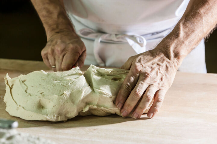 Hände kneten Brotteig auf einer Holzarbeitsfläche, Oberkörper in weißer Schürze erkennbar